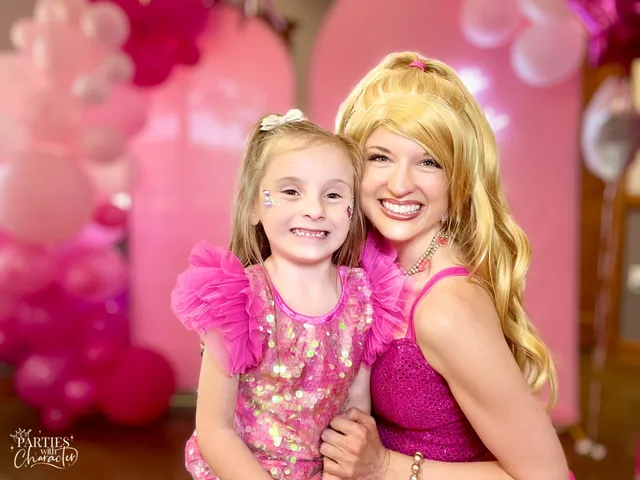 Barbie hugging a young girl in a pink sequin dress beside a balloon arch