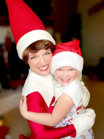 Elf on the Shelf hugging a young girl wearing a matching red Santa hat