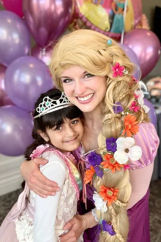 Rapunzel holding a girl in a tiara in front of purple balloons