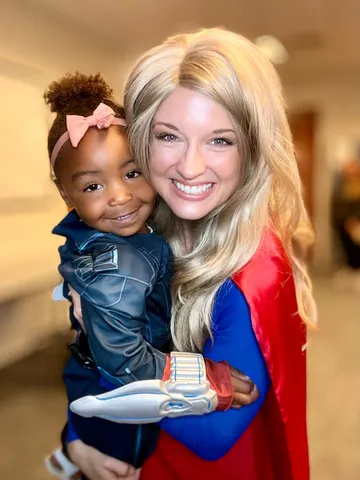 Supergirl carrying a toddler in a superhero suit holding a toy spaceship