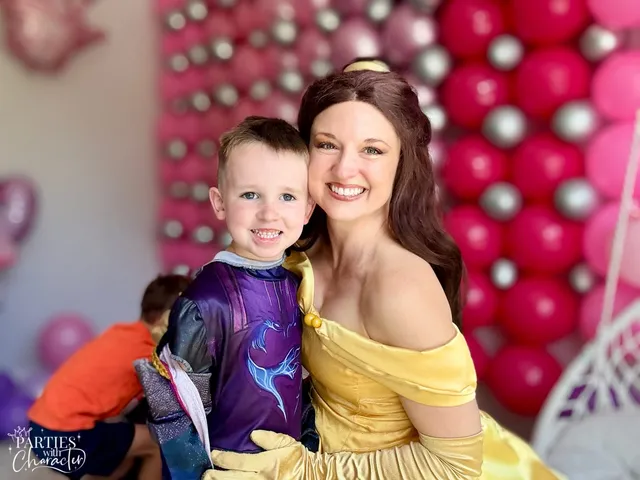 Belle hugging a young boy in a superhero costume against a balloon wall