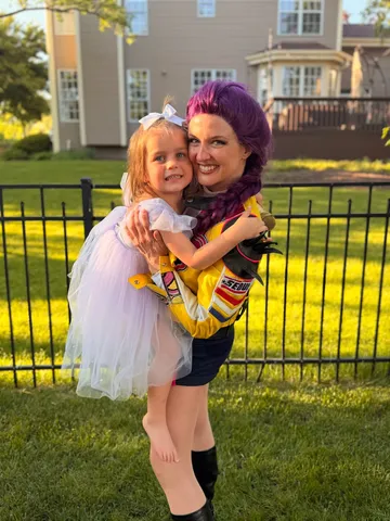 Rumi carrying a toddler in a white tutu at a backyard party