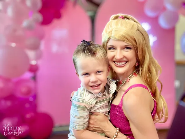 Barbie holding a smiling toddler boy in a striped shirt at a pink party