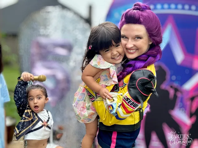 Rumi holding a toddler reaching for a microphone near a bounce house