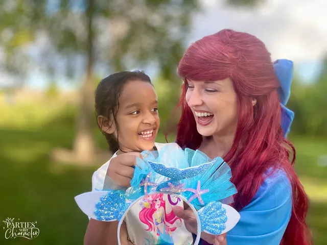 Ariel laughing with a girl holding a mermaid cake topper outdoors