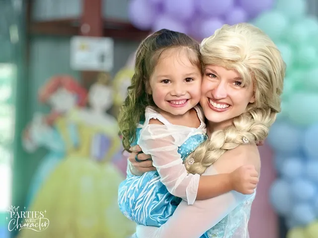 Elsa hugging a girl in a blue gown under a pastel balloon arch at a pavilion