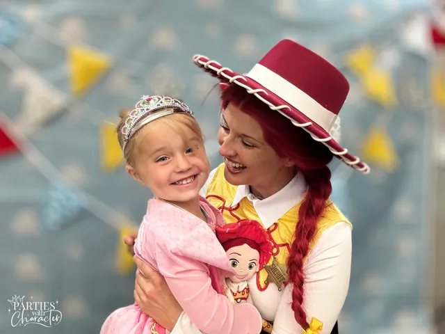 A girl in a pink party dress and tiara held close by Jessie, a Jessie doll tucked between them