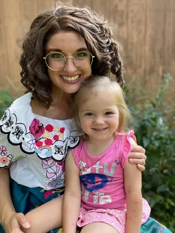 Mirabel holding a toddler in a pink outfit by a wooden fence