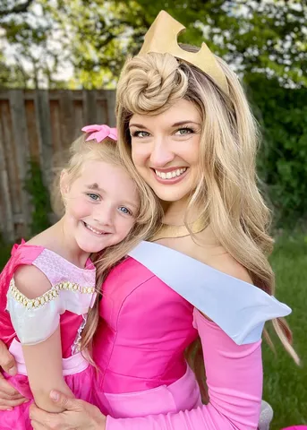 Aurora with a girl in a pink dress and sash in a backyard