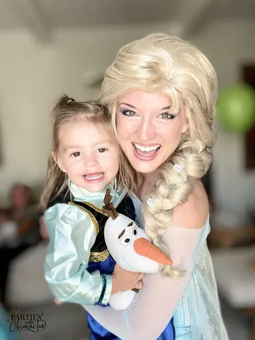 Elsa holding a toddler dressed as Anna clutching an Olaf plush toy indoors
