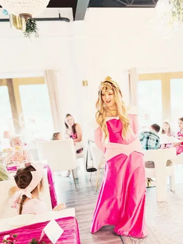 Aurora walking past children seated at pink-draped party tables indoors