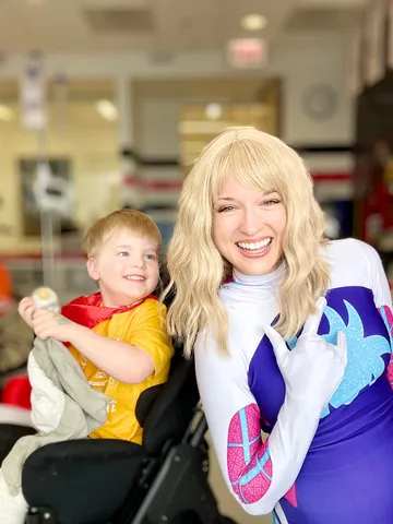 Ghost Spider making a web-shooter sign beside a grinning boy in a wheelchair at a hospital visit