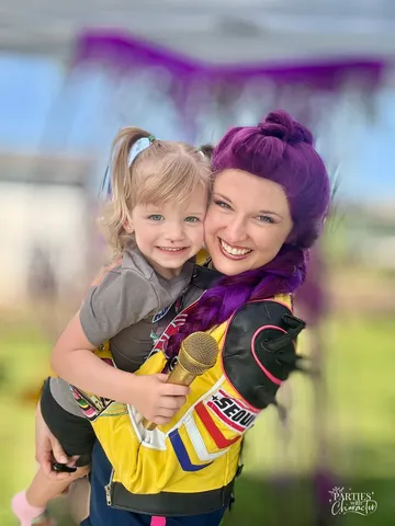 Rumi holding a laughing toddler with a gold microphone under purple streamers