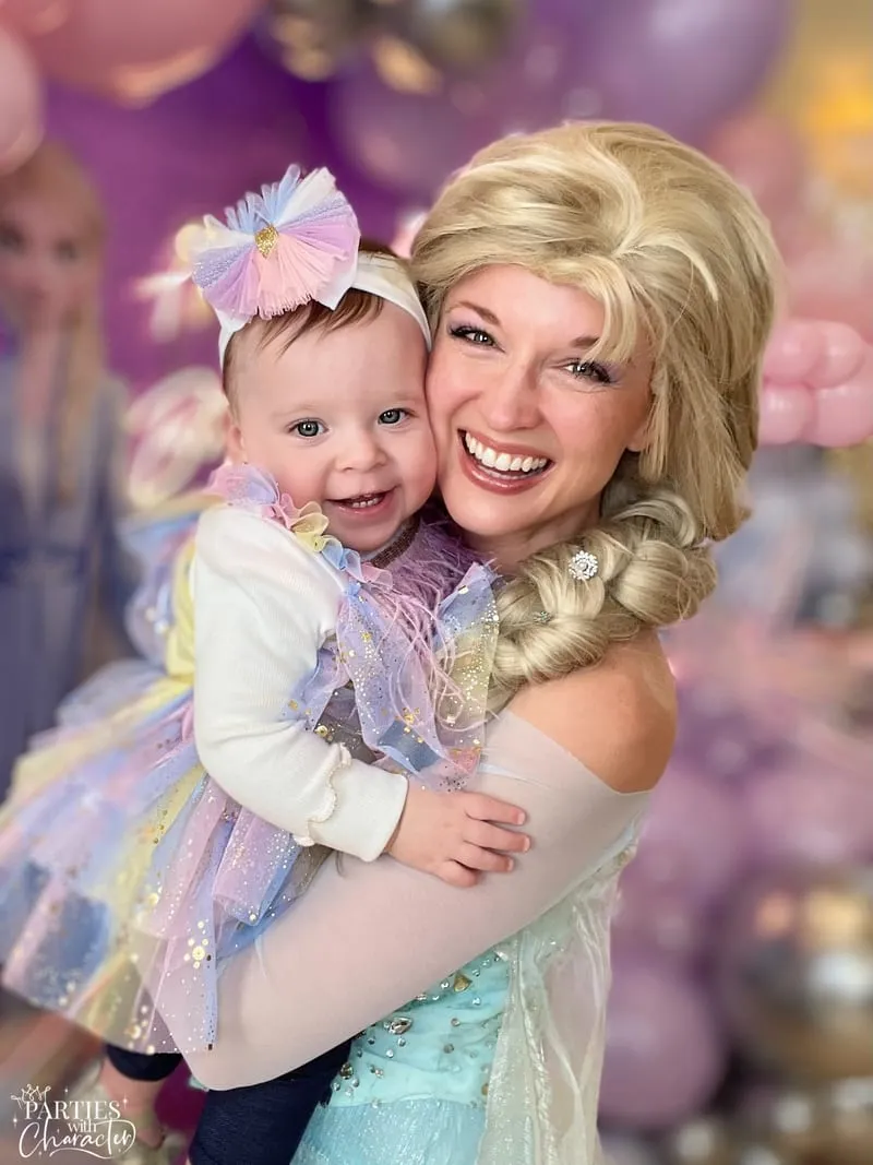 Elsa holding a smiling baby girl in a pastel tutu at a balloon-filled birthday party