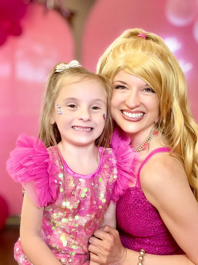 Barbie hugging a young girl in a pink sequin dress beside a balloon arch